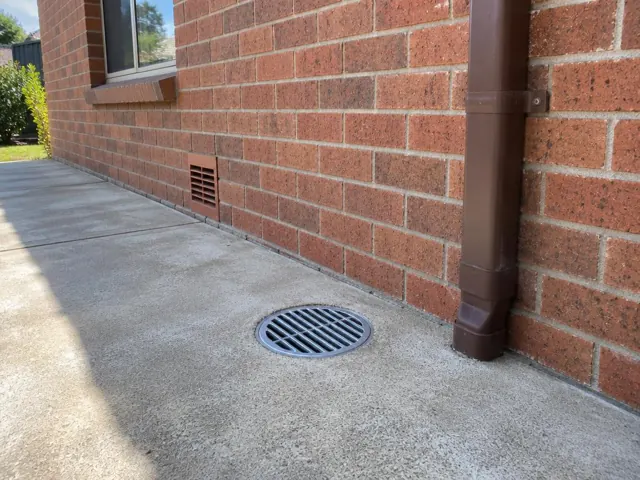 Round metal drain grate beside a brick wall and downpipe on a concrete path in Manly Vale