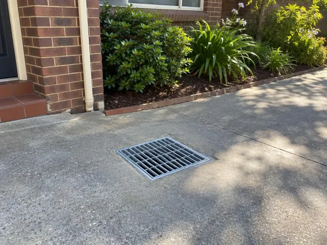Concrete driveway with a flush stormwater grate beside a brick house and garden in Fairlight